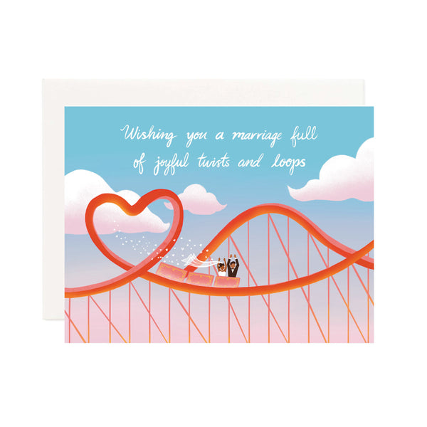 A horizontal wedding card with a scene of a red roller coaster with a heart shaped loop. Has a wedding couple ridding in the coaster car with their arms up and white heart confetti flying behind them. The background has a blue sky with white clouds and a hand lettered saying, "Wishing you a marriage full of joyful twists and loops."